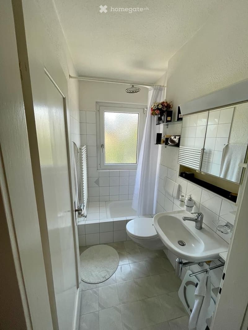 A modern bathroom with white tiles, a bathtub, toilet, sink, mirror, and a shower curtain. A small window provides natural light, and a towel and flower arrangement are placed on the shelf.