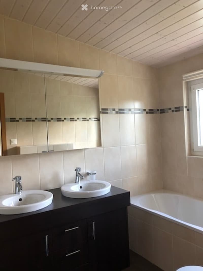 Modern bathroom with double sinks, a large mirror, and a bathtub. The walls are tiled in beige with a black accent border.