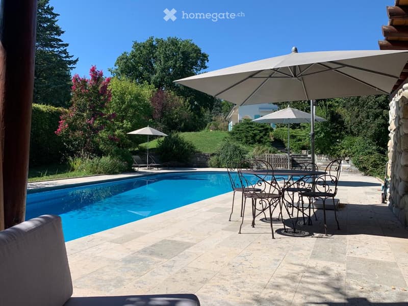 Outdoor patio area with a swimming pool, patio furniture, and umbrellas, surrounded by greenery and trees.