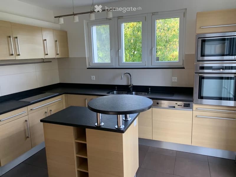Modern kitchen with light wood cabinets, black countertops, a central island, and stainless steel appliances including a microwave and oven. Large windows provide natural light.