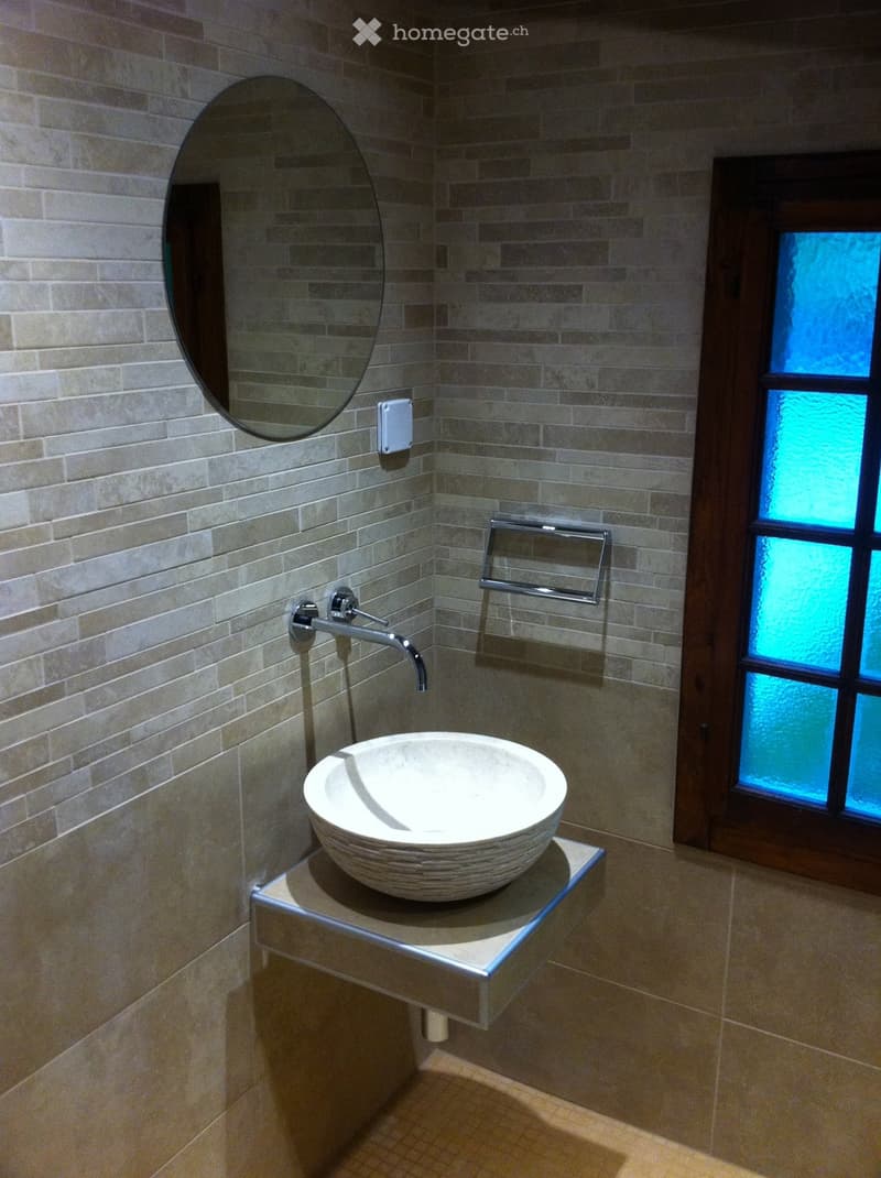 Modern bathroom with a stone sink, chrome faucet, large oval mirror, and towel rack. The walls are tiled in a light beige pattern, and there is a stained-glass window with blue accents.