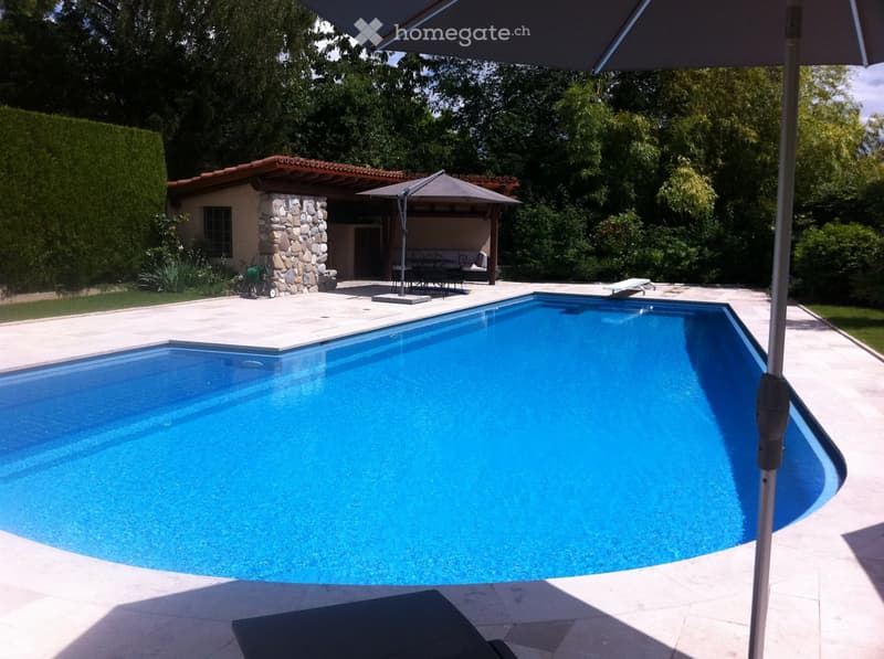A large swimming pool with clear blue water, surrounded by a tiled patio area. A covered outdoor seating area with tables and chairs is visible in the background, along with a stone wall and lush greenery.
