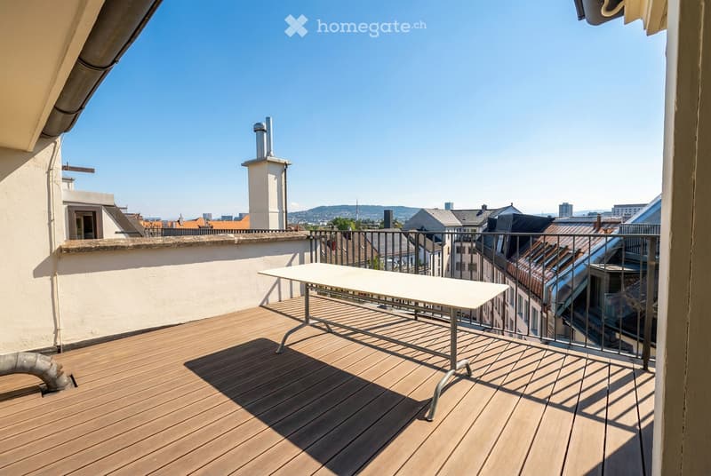 Wooden decked balcony with a white table, metal railing, and cityscape view.