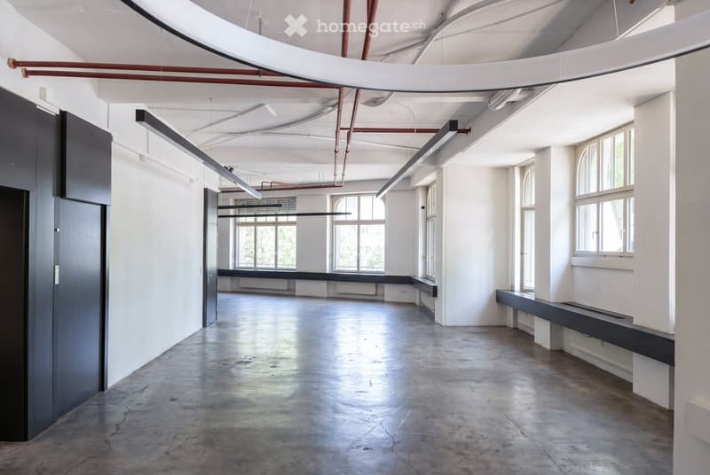 An empty, industrial-style room with white walls, concrete floor, large windows, and exposed pipes and lighting fixtures.