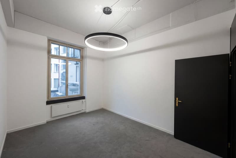 Empty room with white walls, grey carpeted floor, large window with wooden frame, black door, and modern circular pendant light fixture.