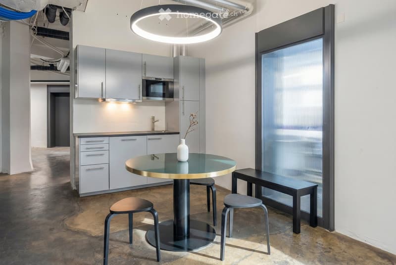 Modern kitchen and dining area with sleek grey cabinets, a round glass table with a gold rim, three stools, a bench, and a vase on the table. The space features exposed pipes and a large circular pendant light.