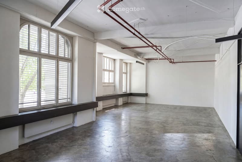 An empty, industrial-style room with concrete floors, white walls, large windows with shutters, and exposed metal pipes on the ceiling.
