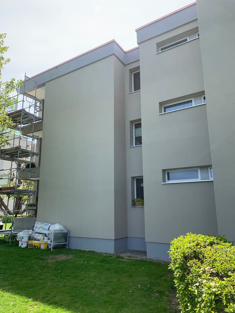 Modern two-story house with light grey exterior, multiple windows, and a small balcony with potted plants. Scaffolding is present on one side, indicating ongoing construction or renovation.