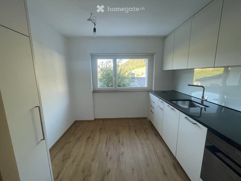 Empty kitchen with white cabinets, wooden flooring, and a window overlooking greenery.