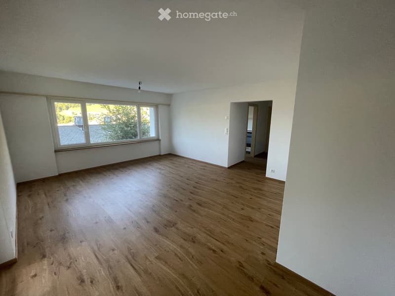 Empty room with light wooden flooring and white walls, featuring a large window with a view of the outdoors and an open doorway leading to another space.