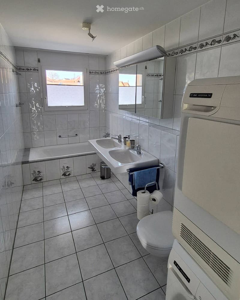 Modern bathroom with a bathtub, double sink, toilet, mirror, and towel rack. Features tiled walls and floor, with a window providing natural light.