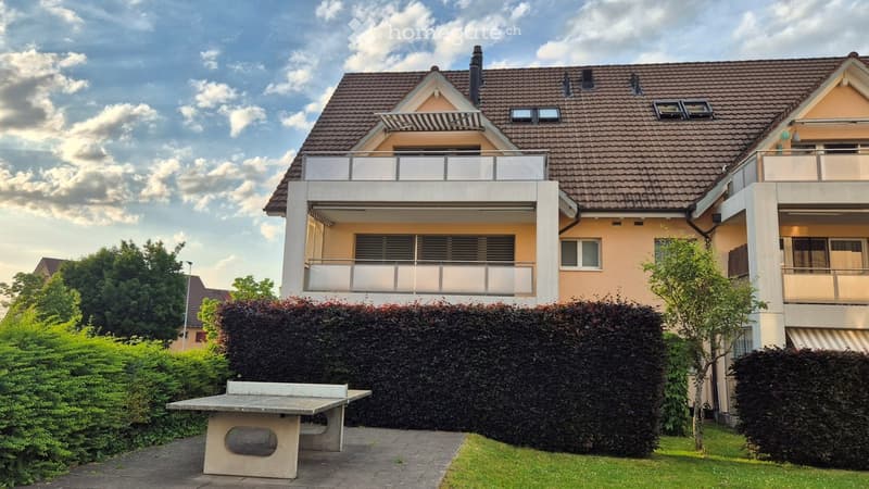 Two-story house with a tiled roof, balconies, and a well-maintained garden featuring a ping pong table and trimmed hedges.