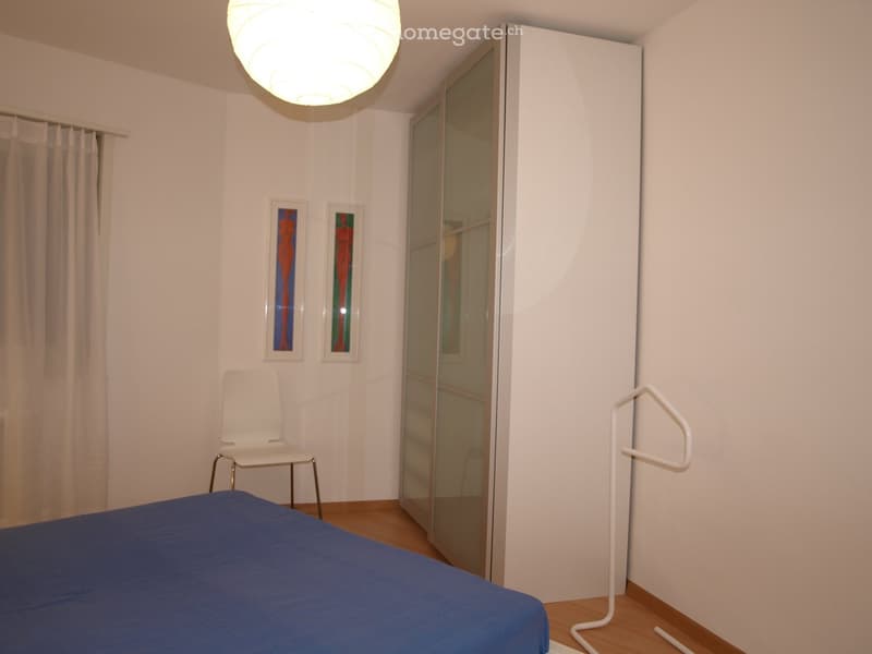 Minimalist bedroom with a blue bedspread, white chair, large mirror, and two framed artworks on the wall.