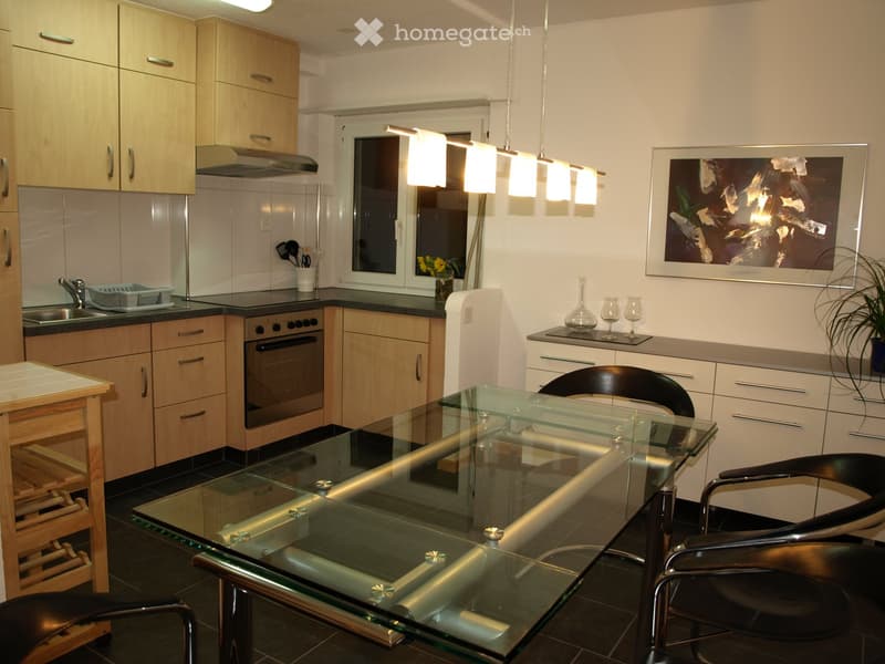 Modern kitchen with light wooden cabinets, stainless steel appliances, a glass dining table with black chairs, and a vase with wine glasses on the counter.