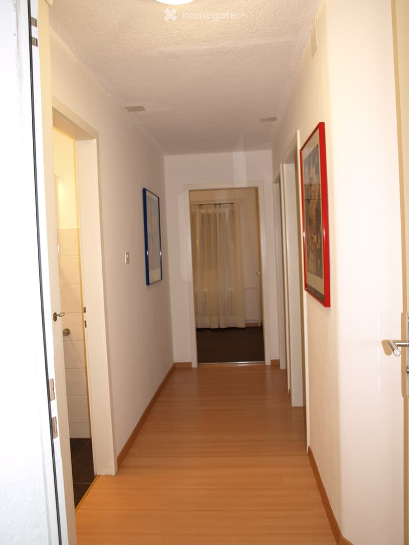 A clean, well-lit hallway with wooden flooring, white walls, and several doors leading to different rooms. Two framed pictures are displayed on the walls.