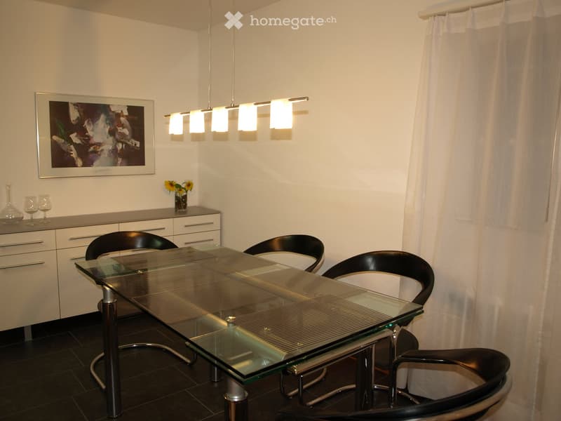 Modern dining area with a sleek glass table, black chairs, a white cabinet, and a pendant light fixture.