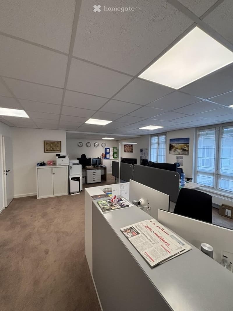 Office space with desks, chairs, computer screens, cabinets, and a printer. Large windows provide natural light.
