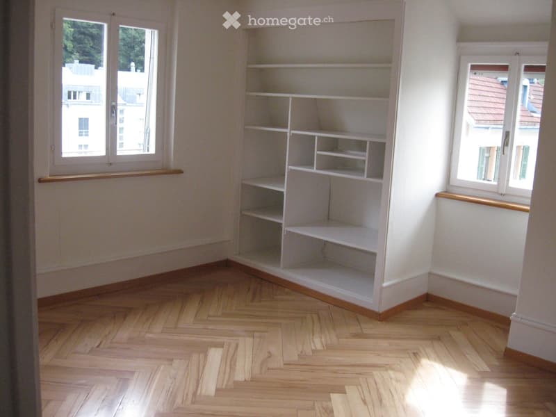 Empty room with wooden herringbone flooring, white walls, built-in shelving unit, and large windows with wooden sills.