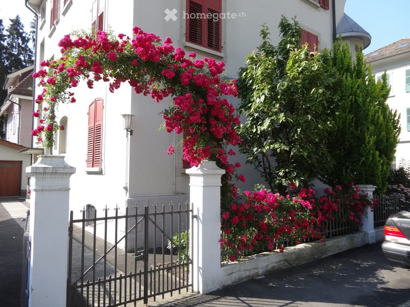 A white house with red shutters, surrounded by lush greenery and vibrant pink flowers climbing over the facade. A black wrought-iron fence and white pillars frame the entrance.