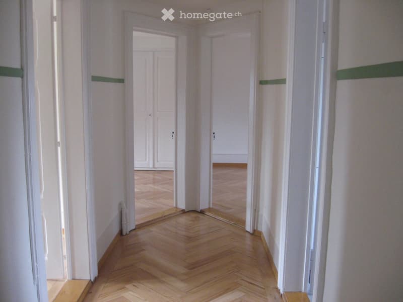 Empty hallway with wooden flooring and white walls, featuring multiple doors leading to other rooms.