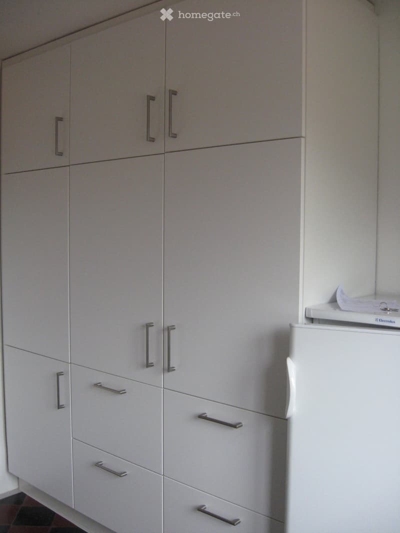 Modern kitchen with white cabinets and a stainless steel refrigerator. The cabinets have sleek, minimalist handles and are arranged in a vertical stack.