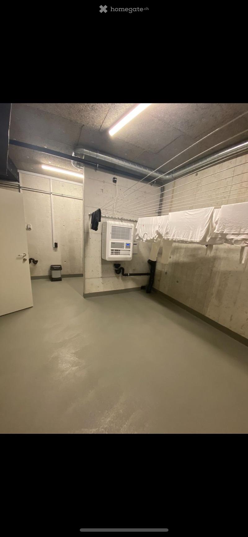 Empty utility room with concrete walls and floor, fluorescent lighting, a clothes drying rack, a small sink, and a white air conditioning unit.