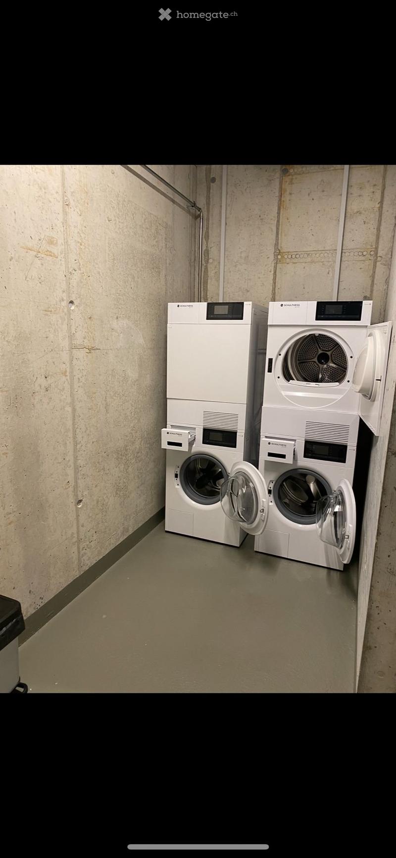 A small utility room with a washing machine and dryer, both white and modern, placed side by side against a concrete wall.