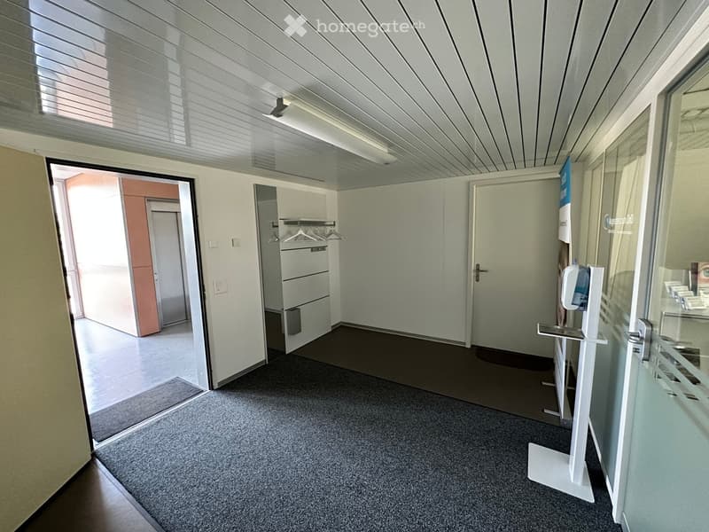 Empty hallway with grey carpet, white walls, and a ceiling with recessed lighting. Features include a coat rack, a hand sanitizer dispenser, and an open doorway leading to another room.