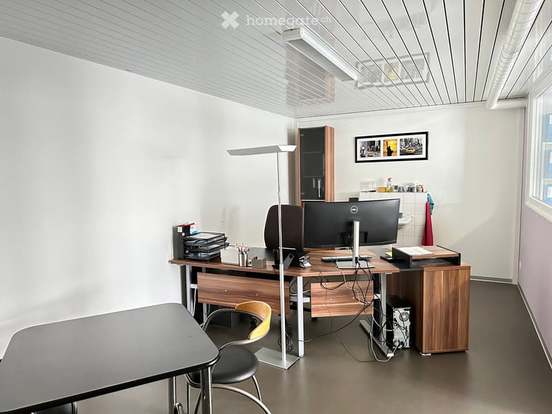 Modern office space with a wooden desk, ergonomic chair, computer setup, floor lamp, and a small meeting table.