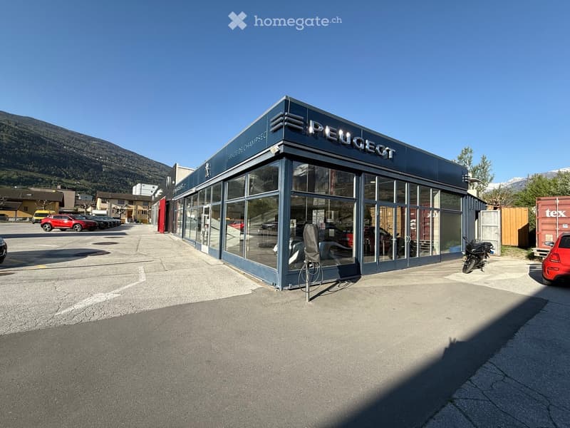 A modern Peugeot car dealership with large glass windows and a flat roof, situated in a parking lot with several cars and motorcycles visible.