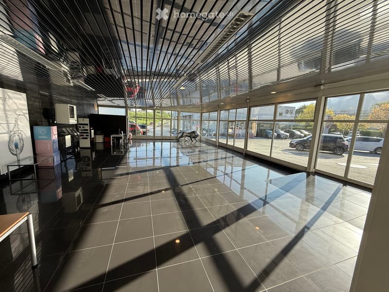 Modern, spacious hallway with large glass windows and a sleek, reflective tiled floor. The ceiling features a geometric, angular design with integrated lighting. Visible outside are parked cars and a green landscape.
