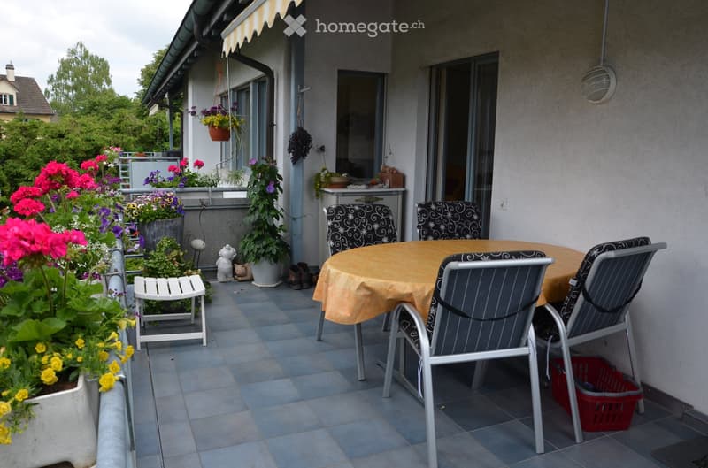 Balcony with tiled floor, round table with yellow tablecloth, four chairs, small white stool, flower pots, and a small toy figure.