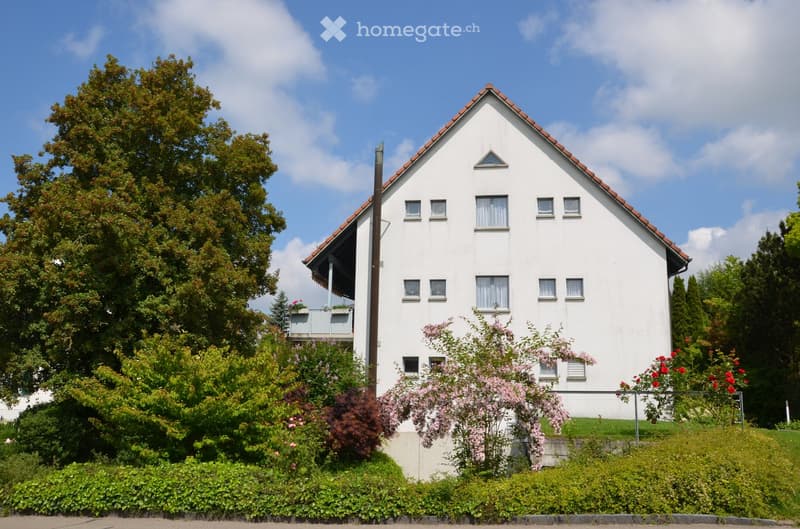 Two-story white house with red-tiled roof, multiple windows, and a small balcony on the upper floor. Surrounded by lush greenery and flowering shrubs.