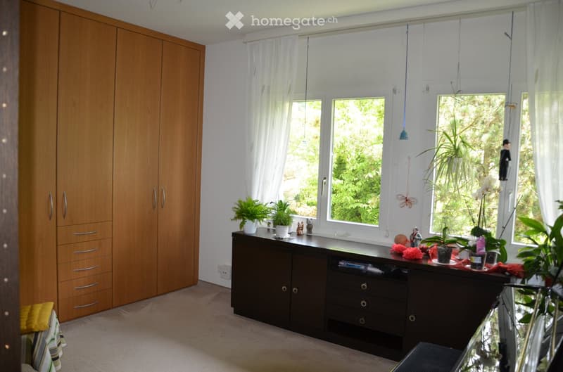 A well-lit room with large windows, a wooden wardrobe, a dark wooden cabinet with drawers, and several potted plants on the windowsill.