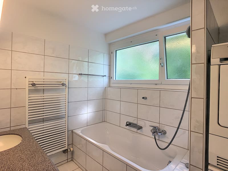 Modern bathroom with a bathtub, sink, radiator, and window with frosted glass. The walls are tiled in white and beige.