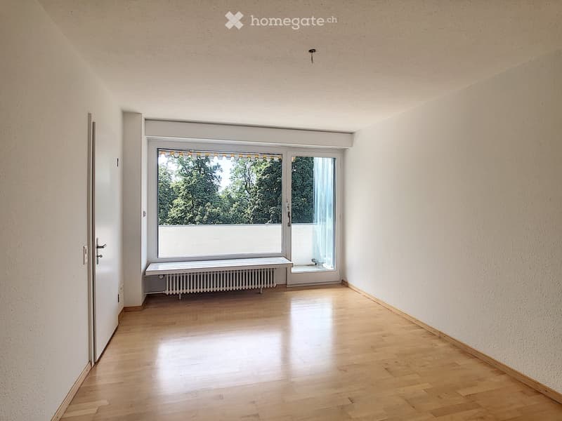 Empty room with wooden flooring, white walls, large window with a bench, and a door leading to a balcony.