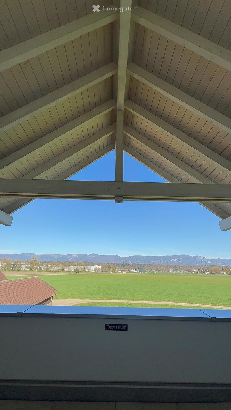 Balcony with wooden ceiling beams and open sides, overlooking a green field and distant mountains.