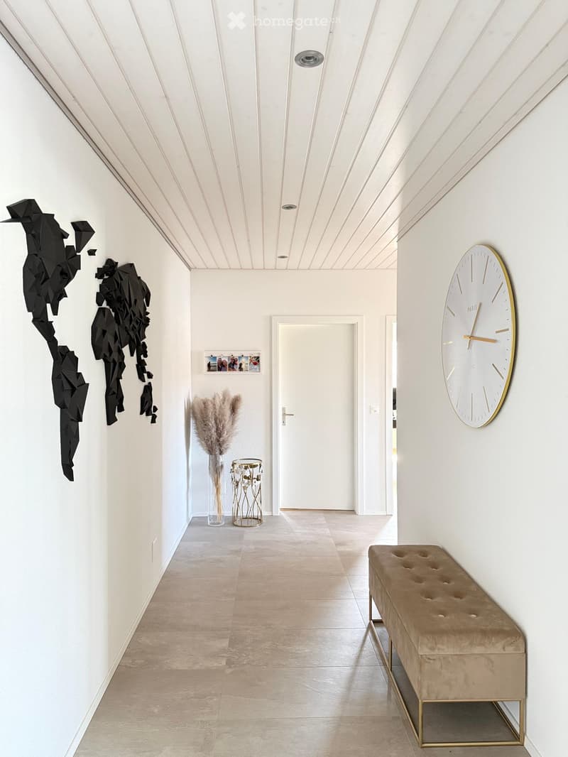 Modern hallway with white walls, geometric art, a large clock, a bench, and a vase with dried plants.