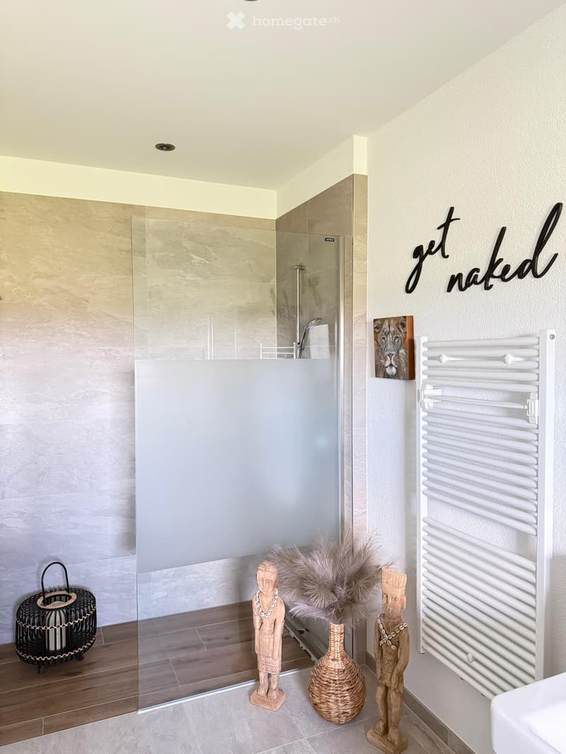 Modern bathroom with a glass-enclosed shower, a white radiator, decorative statues, a wicker basket, and a vase with feathers.