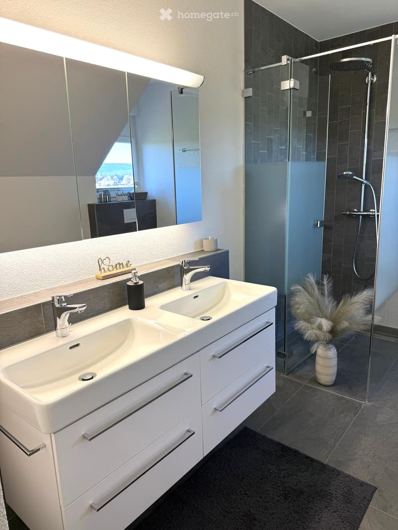 Modern bathroom with double sink, large mirror, glass-enclosed shower, and decorative vase with feathers.