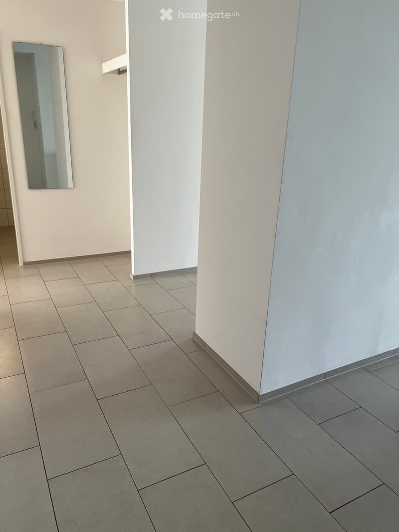 Empty hallway with tiled floor and white walls, featuring a mirror on one wall and a small shelf.