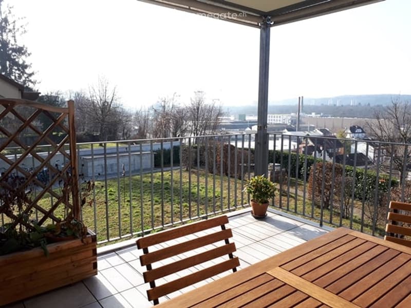 Balcony with wooden table and chairs, potted plants, metal railing, and a view of the city and greenery.