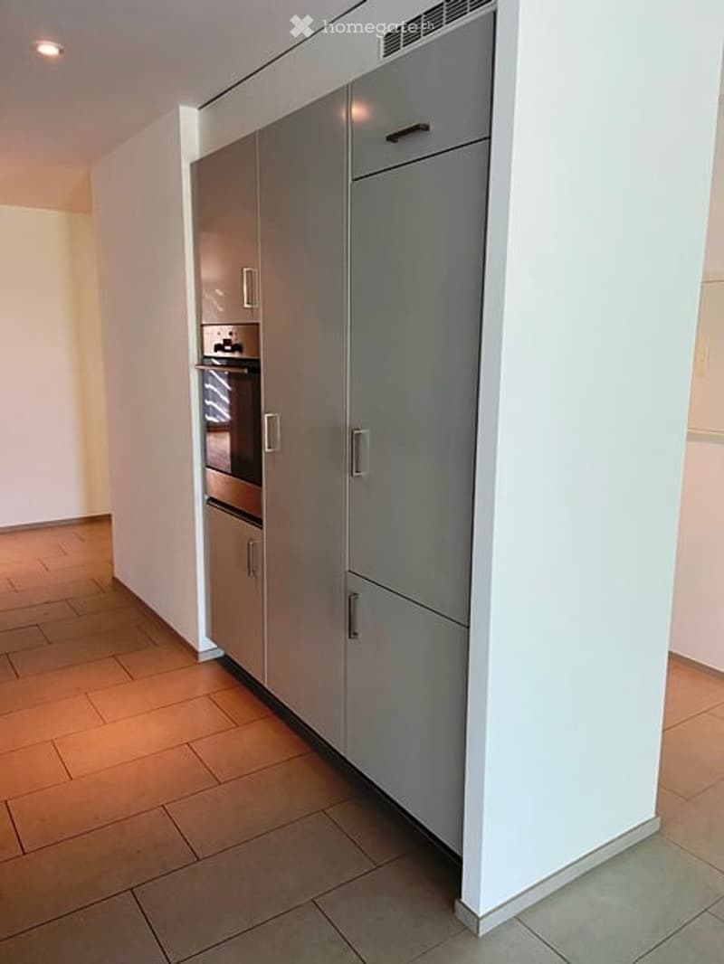 Modern kitchen with stainless steel appliances, including a refrigerator and oven, set against white walls and a wooden floor.