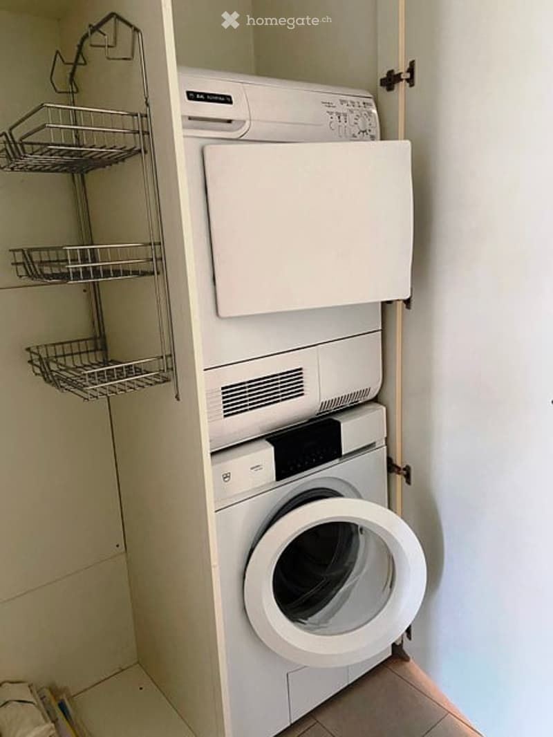 Compact bathroom with a stacked washing machine and dryer unit, metal shelf above, white tiled walls and floor.