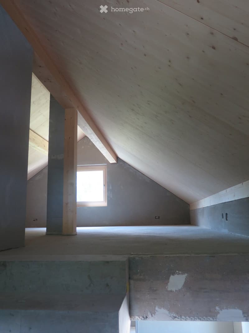 Empty attic space with wooden beams, concrete floor, and a small window letting in natural light. No furniture or furnishings present.