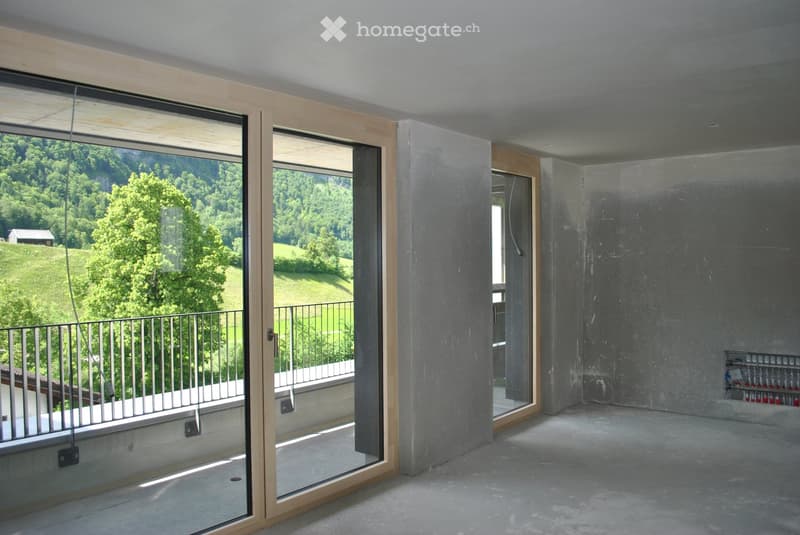 Empty room with large glass windows and a balcony overlooking a green landscape. The room features concrete floors and walls, with a metal railing on the balcony.