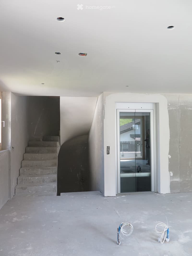 Empty room with white walls, concrete floor, and a staircase leading to another level. A glass elevator door is visible on the right side.