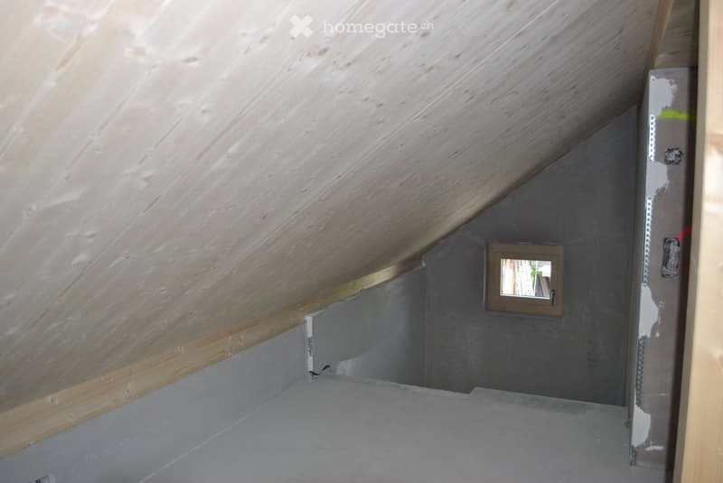 Empty attic space with wooden slanted ceilings, grey walls, and a small window with a brown frame.