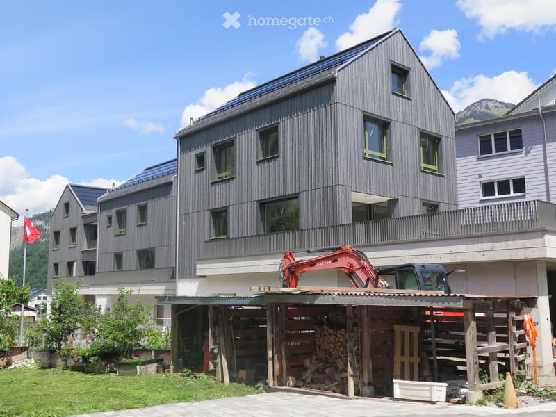 Modern multi-story building with dark wooden facade and green-framed windows, featuring a balcony on the upper level. A construction vehicle is parked under a covered area with stacked firewood and tools.
