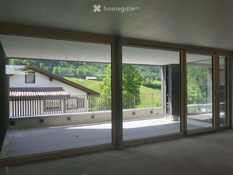 An unfurnished room with large sliding glass doors opening to a balcony, overlooking a green landscape with a house and trees.